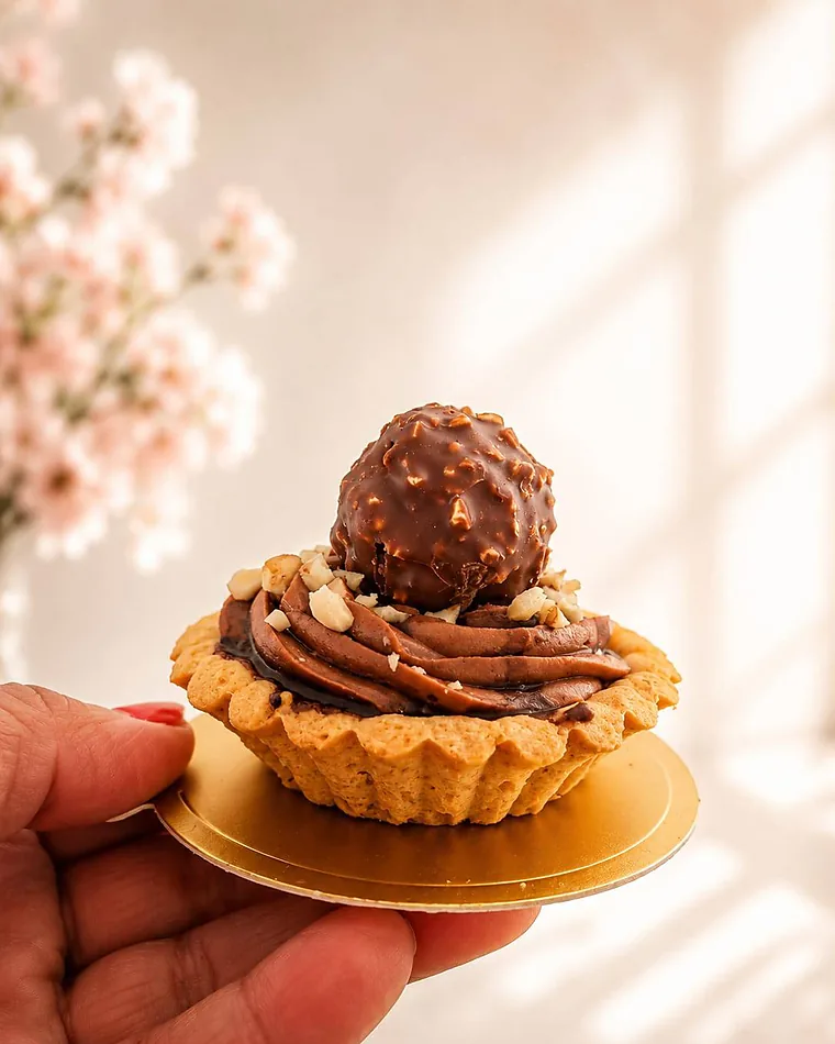 A hand holding a golden chocolate hazelnut tartlet crowned with a whole Ferrero Rocher